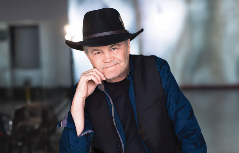 Promotional headshot portrait of Micky Dolenz of The Monkees wearing a black fedora, dark vest, and blue shirt resting his chin on his hand.